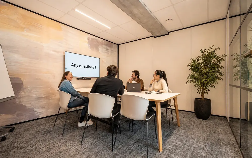 Quatre personnes échangent autour d'une table, devant un écran affichant « Any questions ? »
